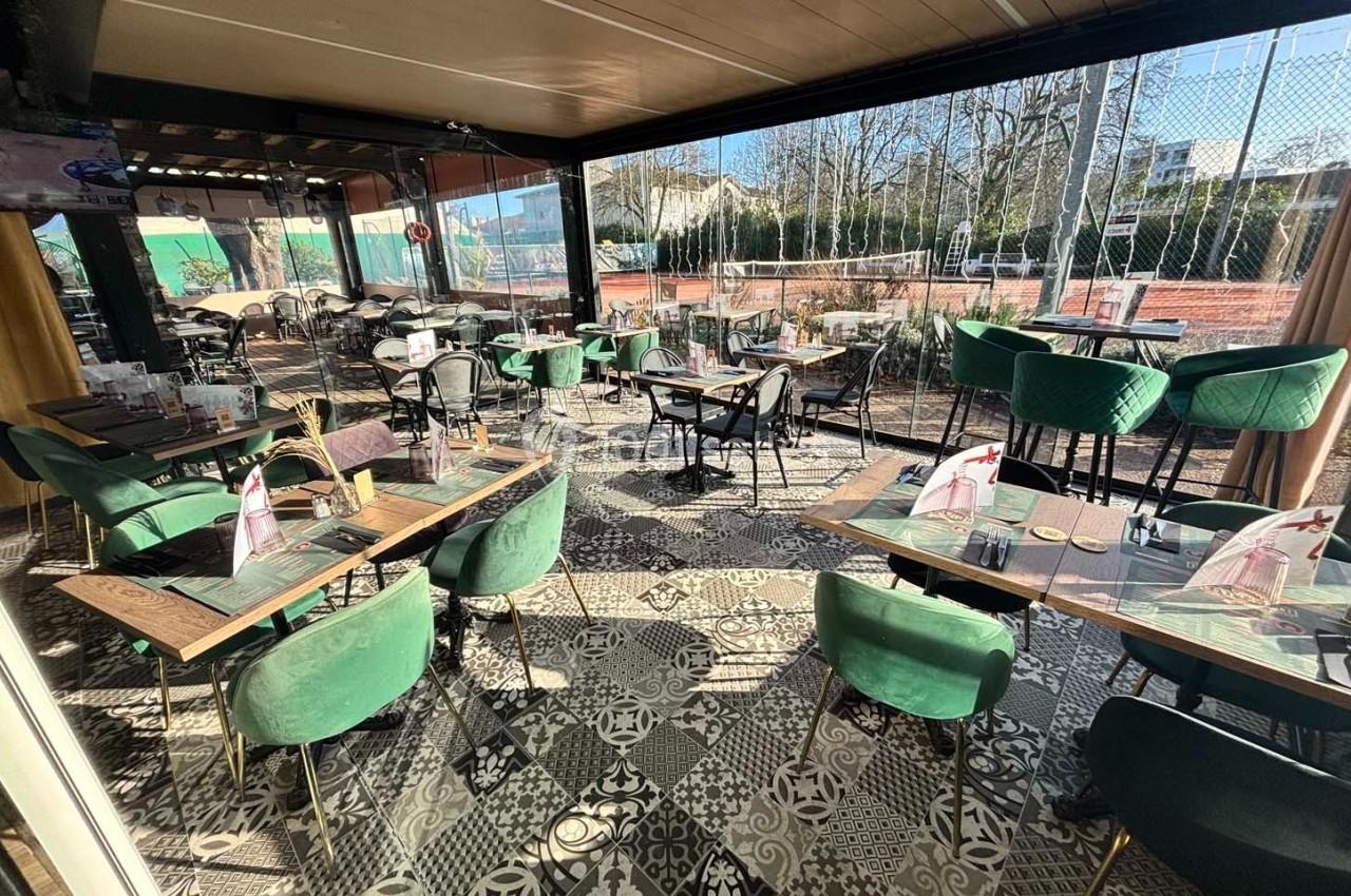 Salle de restaurant lumineuse avec tables et chaises vertes, vue sur des terrains de tennis à l'extérieur.