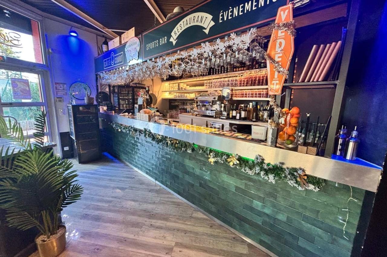 Comptoir de bar décoré avec des guirlandes, étagères remplies de verres et bouteilles, ambiance lumineuse et festive.