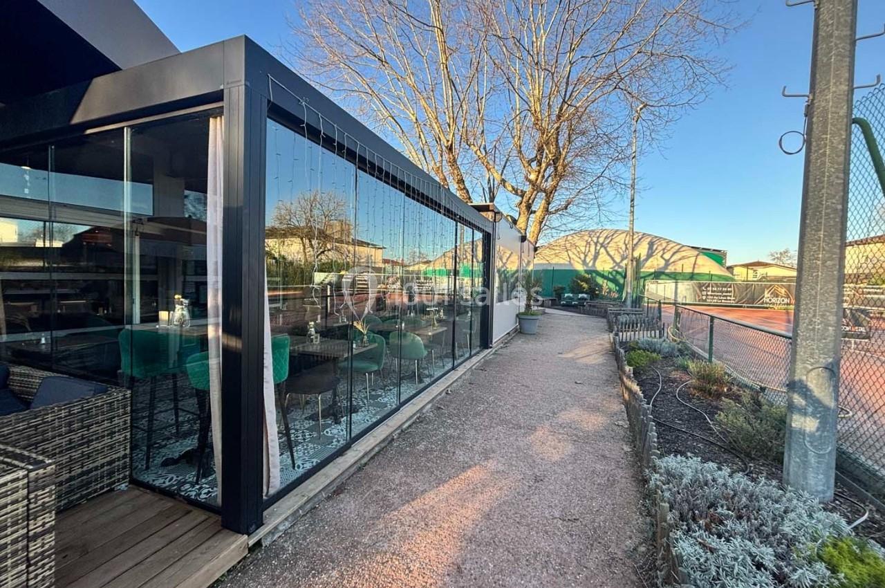 Terrasse vitrée moderne donnant sur un chemin en gravier, avec des terrains de tennis en arrière-plan.