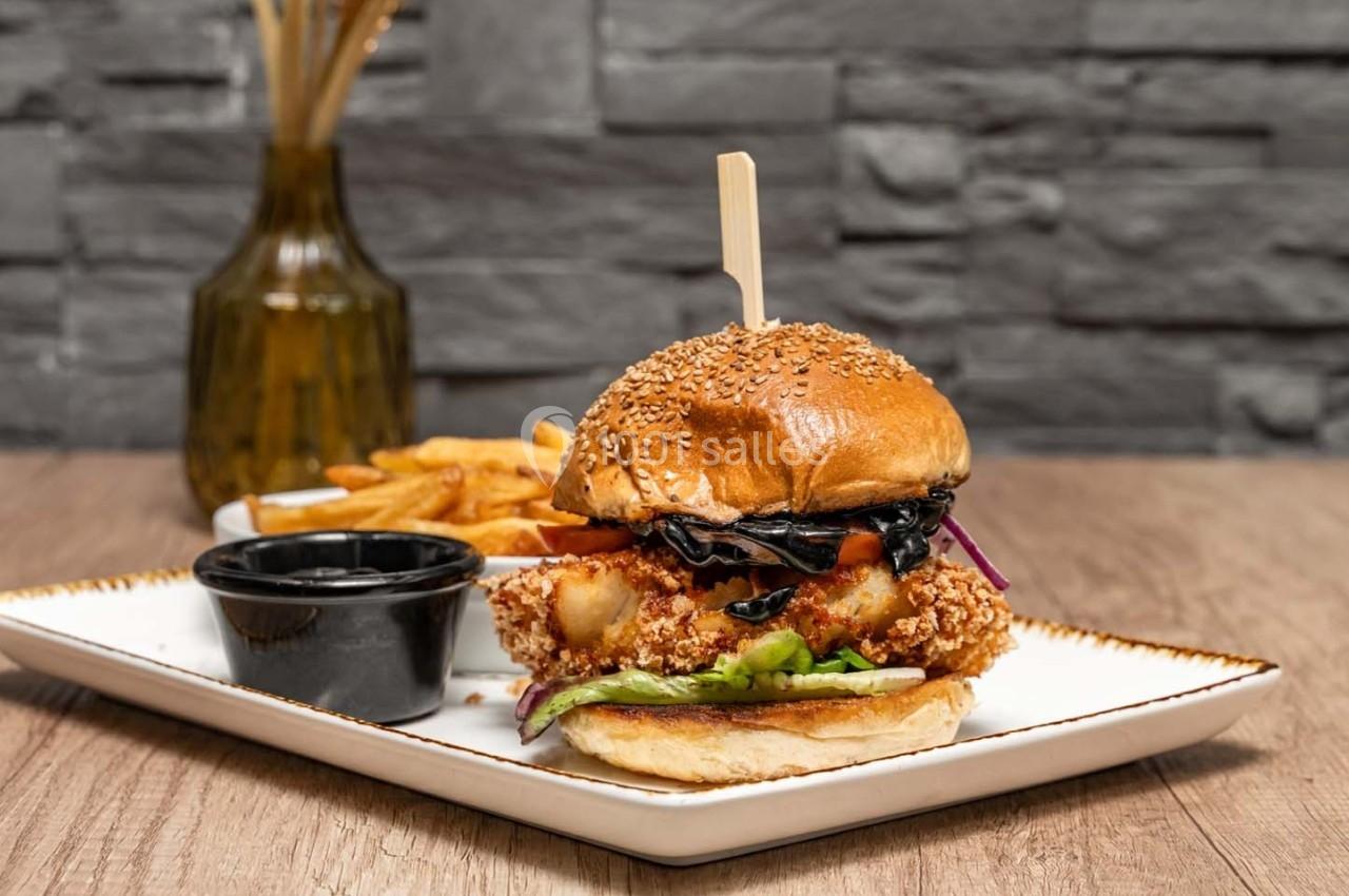 Burger au poulet pané garni de légumes, servi avec des frites et une sauce, présenté sur une assiette blanche.