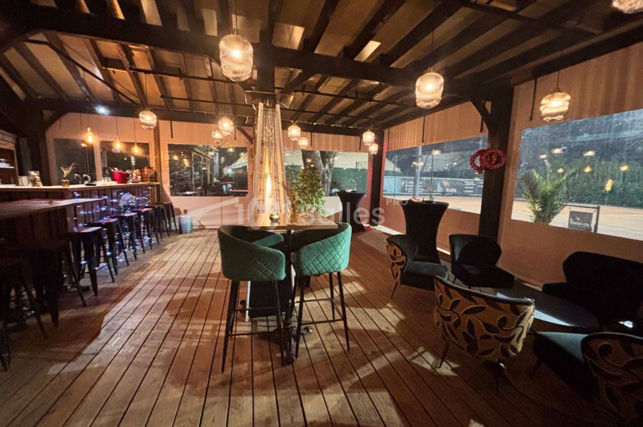 Terrasse couverte en bois avec tables, chaises et éclairage suspendu, ambiance chaleureuse en soirée.