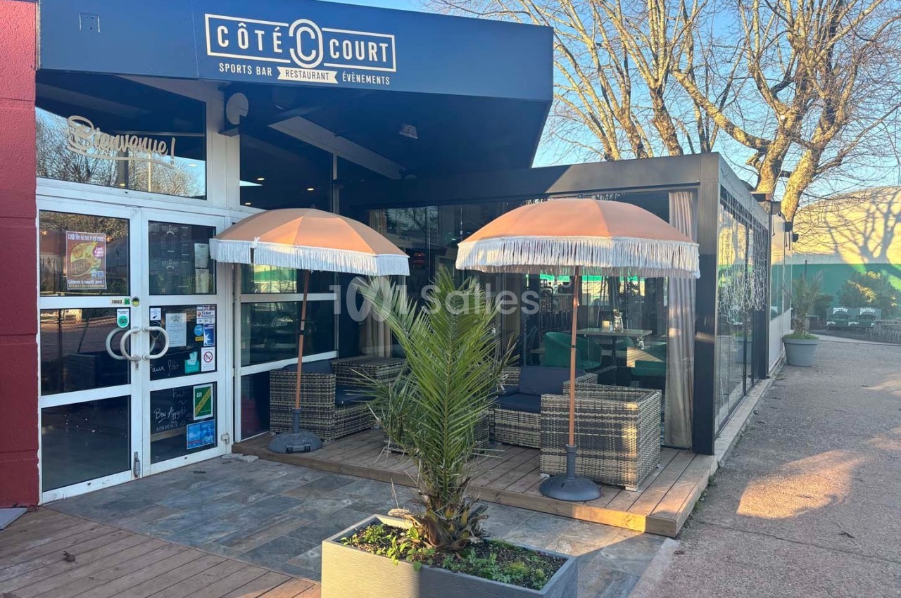 Terrasse d'un restaurant avec parasols, mobilier en rotin et végétation, située devant une entrée vitrée.
