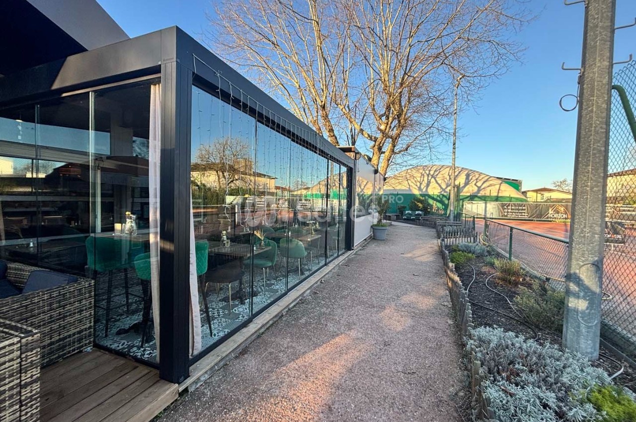 Terrasse vitrée d'un restaurant donnant sur un chemin en gravier, avec vue sur des terrains de tennis et un arbre dénudé.