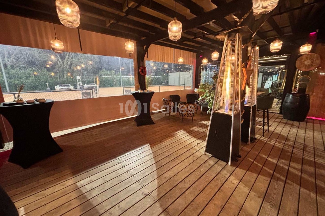 Terrasse en bois couverte, éclairée par des suspensions lumineuses, avec tables hautes et chauffages d'extérieur.