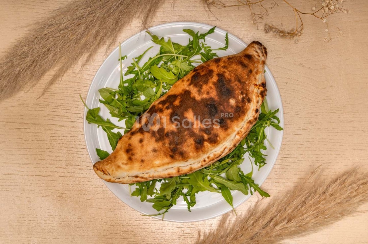 Calzone doré servi sur un lit de roquette dans une assiette blanche, posé sur une table en bois clair avec des décorations…