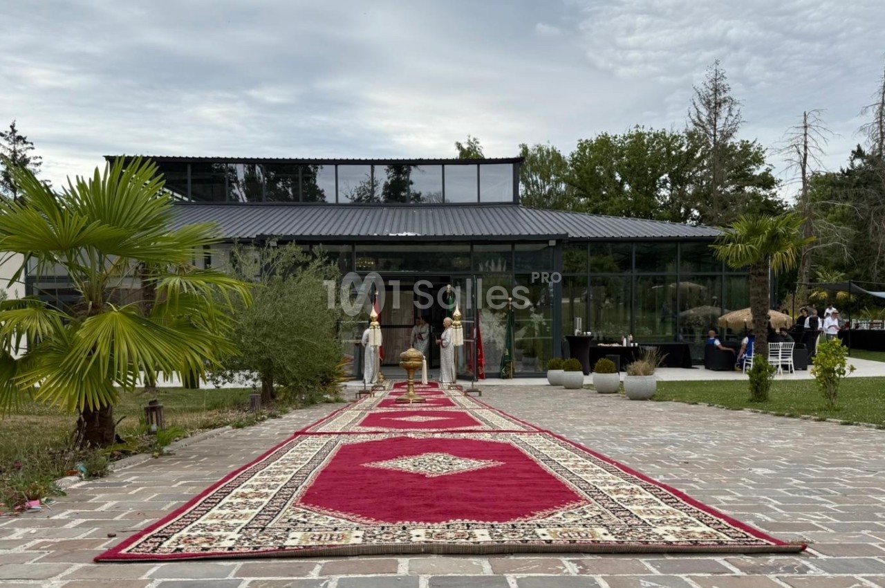 Allée en pavés bordée de palmiers menant à une verrière, avec tapis rouges ornés de motifs géométriques.