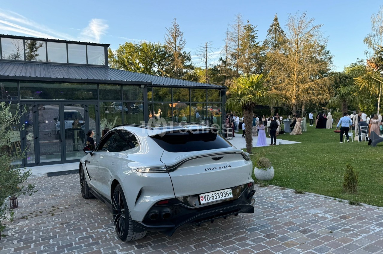 Voiture grise garée devant un bâtiment vitré, avec des invités rassemblés dans un jardin à l'arrière-plan.
