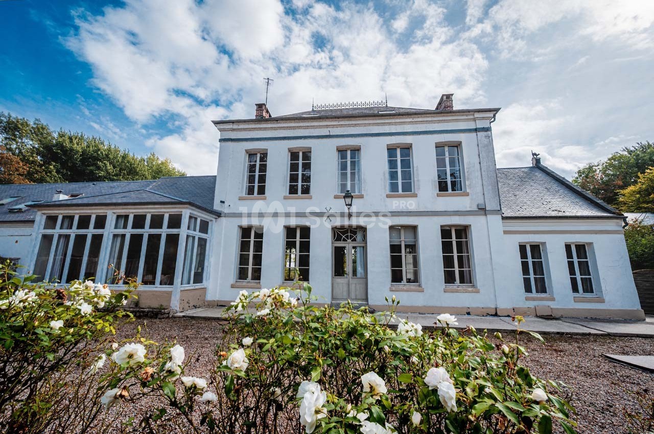 Façade d'une maison blanche à deux étages avec des fenêtres symétriques, entourée de rosiers en fleurs.