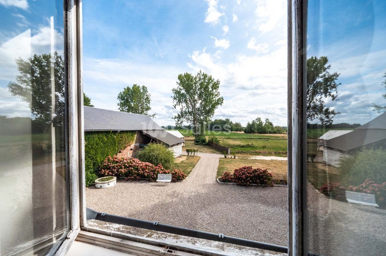 Vue depuis une fenêtre ouverte sur une cour gravillonnée, entourée de bâtiments et de végétation, sous un ciel partiellement…