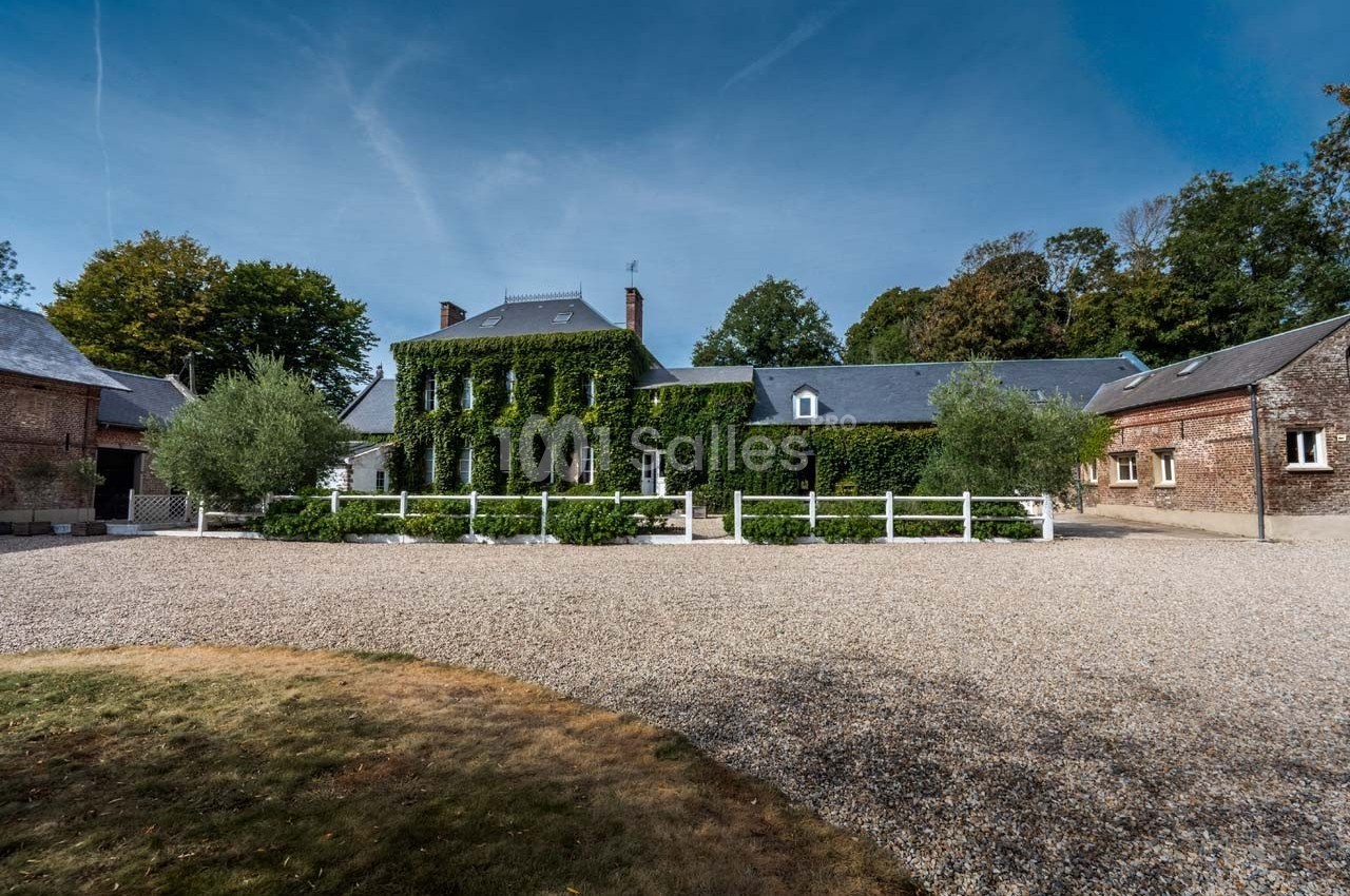 Grande maison en pierre recouverte de lierre, entourée d'arbres, avec une cour en gravier au premier plan.