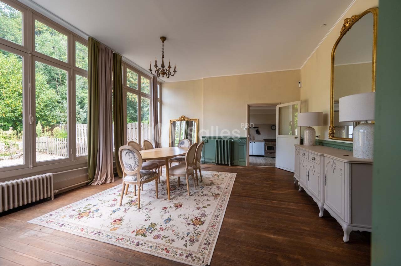 Salle à manger lumineuse avec table en bois, chaises élégantes, tapis fleuri, miroir doré et grandes fenêtres donnant sur un…
