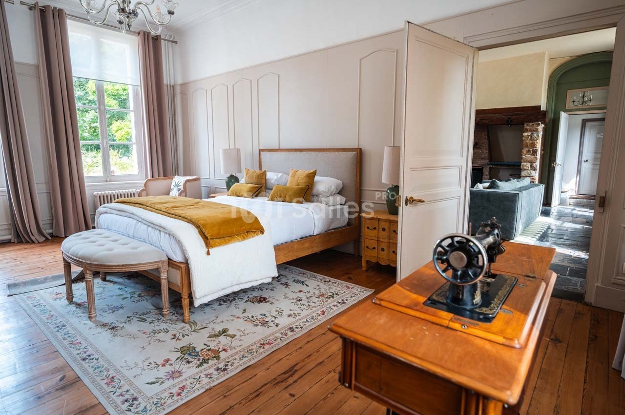 Chambre lumineuse avec lit double, coussins jaunes, tapis floral, machine à coudre ancienne et vue sur un salon en arrière…