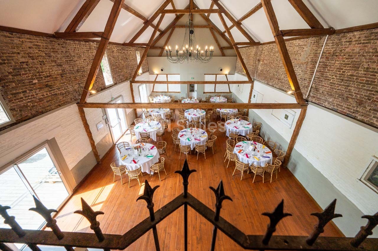 Salle de réception avec charpente en bois apparente, tables rondes dressées et sol en parquet.