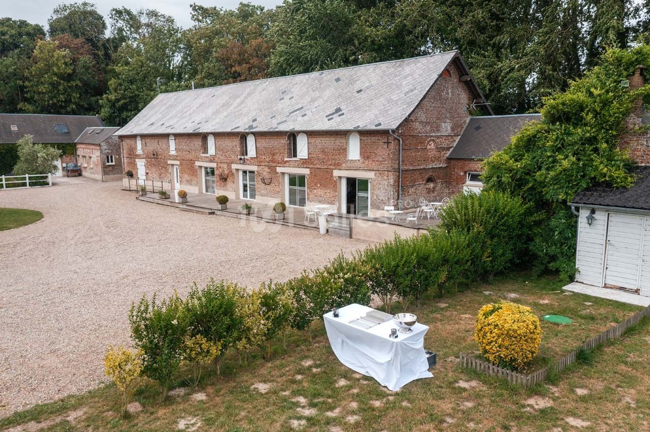 Bâtiment en briques avec toit en ardoise, entouré de végétation et d'une cour gravillonnée, table dressée au premier plan.
