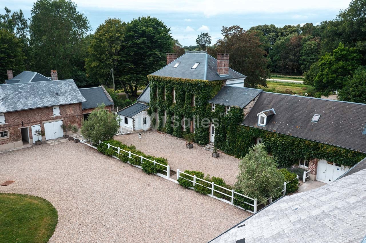 Vue aérienne d'une grande maison en briques recouverte de lierre, entourée d'une cour gravillonnée et de bâtiments annexes.
