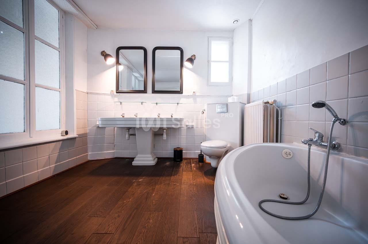 Salle de bain lumineuse avec baignoire, double vasque, carrelage blanc, parquet foncé et fenêtres vitrées.