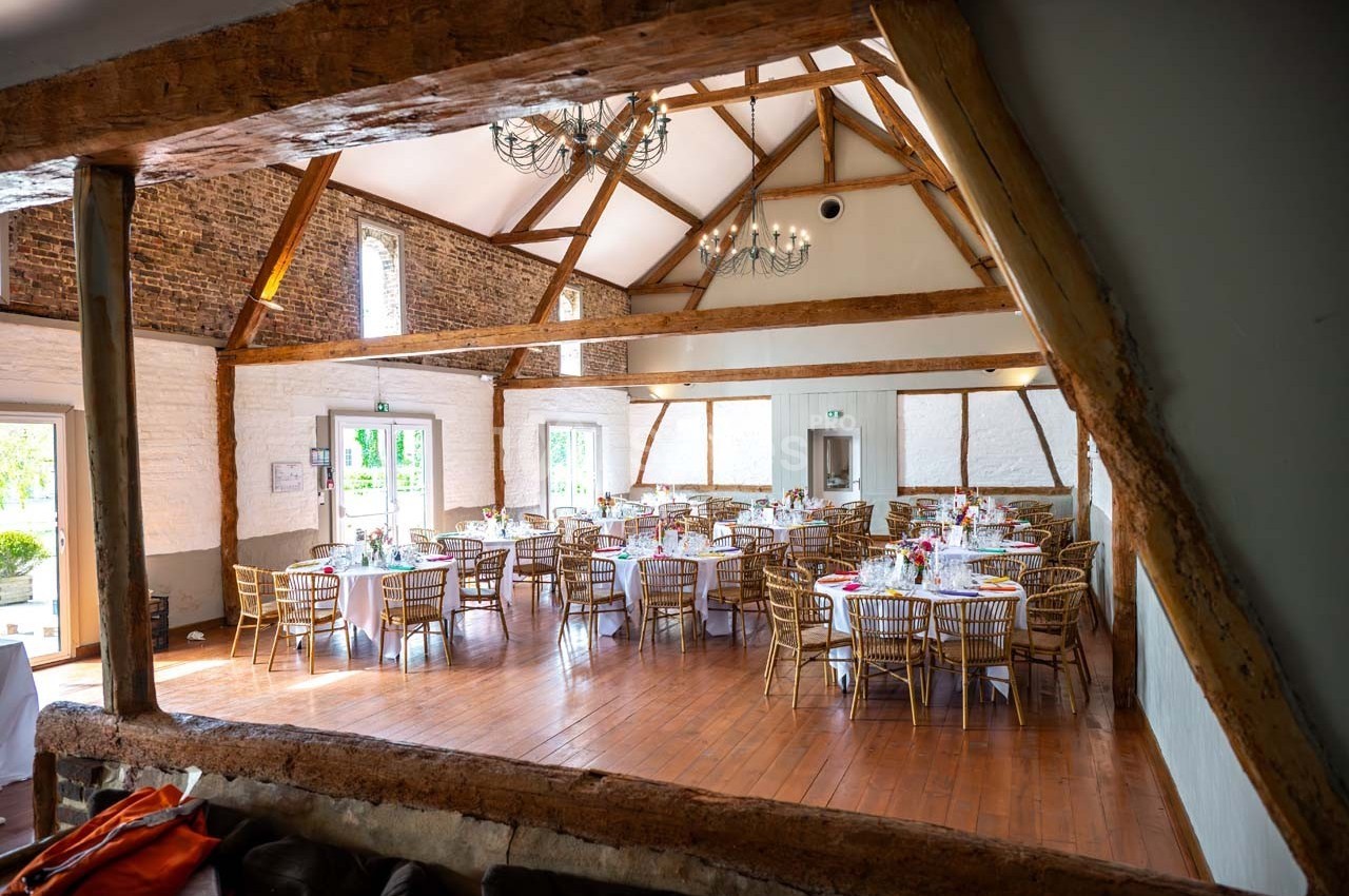 Salle de réception lumineuse avec poutres apparentes, tables rondes dressées et chaises en bois.