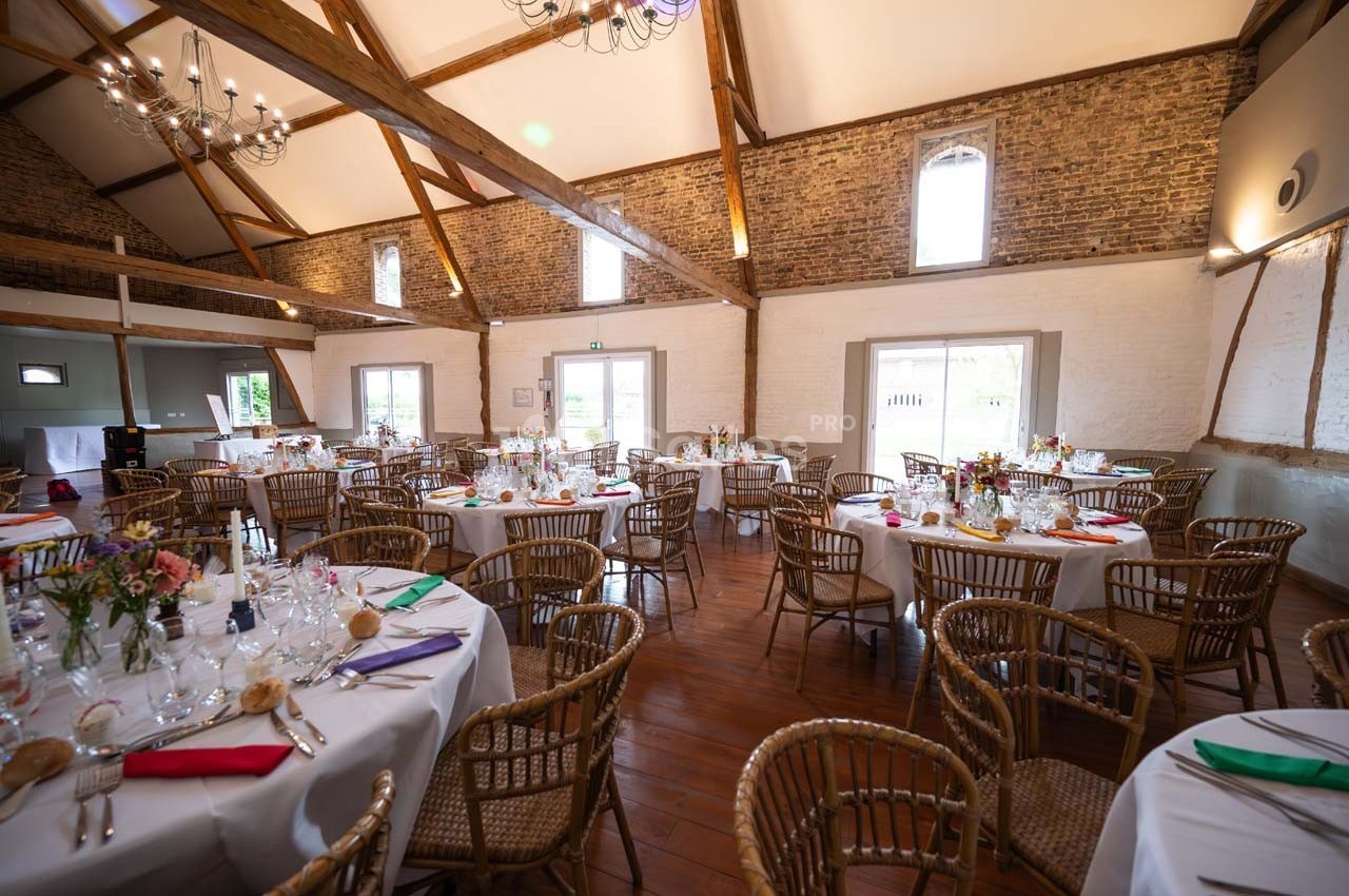 Salle de réception lumineuse avec murs en briques, poutres apparentes et tables rondes décorées pour un événement.