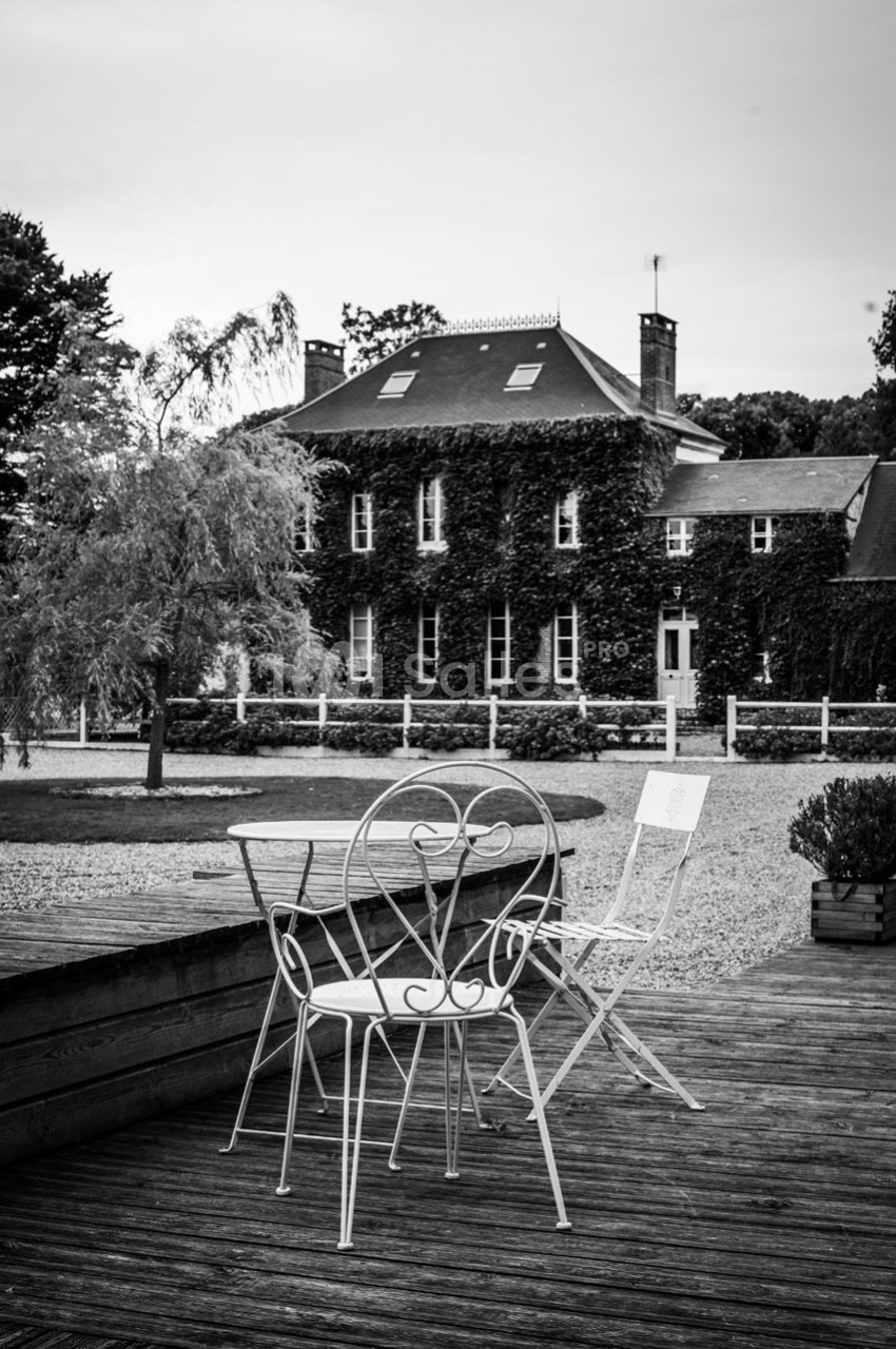 Chaises et table en fer forgé sur une terrasse en bois, avec une maison couverte de lierre en arrière-plan.