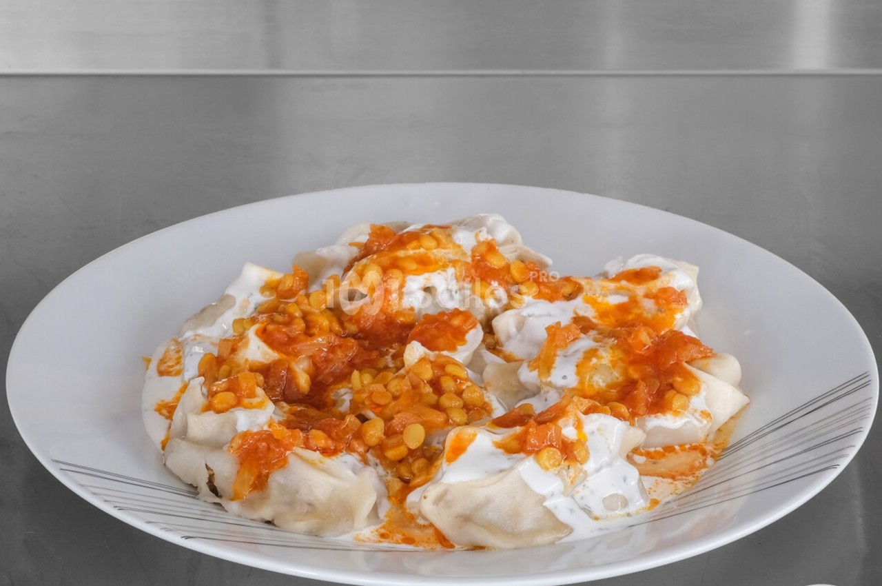 Assiette de raviolis garnis de yaourt, sauce tomate et pois cassés, présentée sur une table en acier inoxydable.