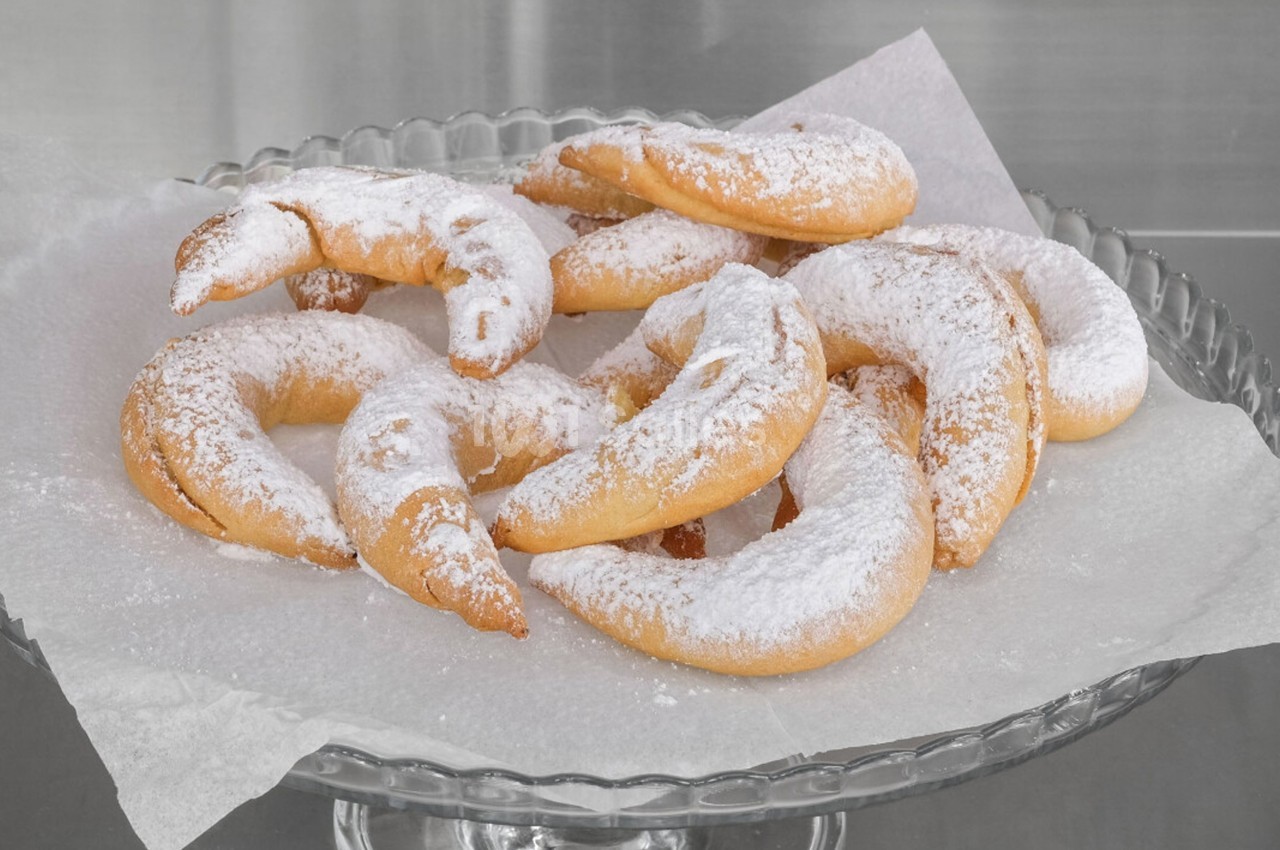 Assortiment de croissants sablés saupoudrés de sucre glace, disposés sur un plat en verre avec du papier cuisson.