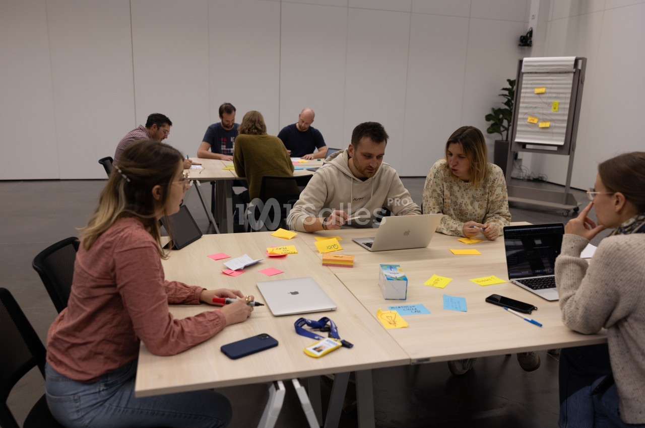 Des personnes travaillent en groupe autour de tables avec des ordinateurs portables, des notes adhésives et des documents.
