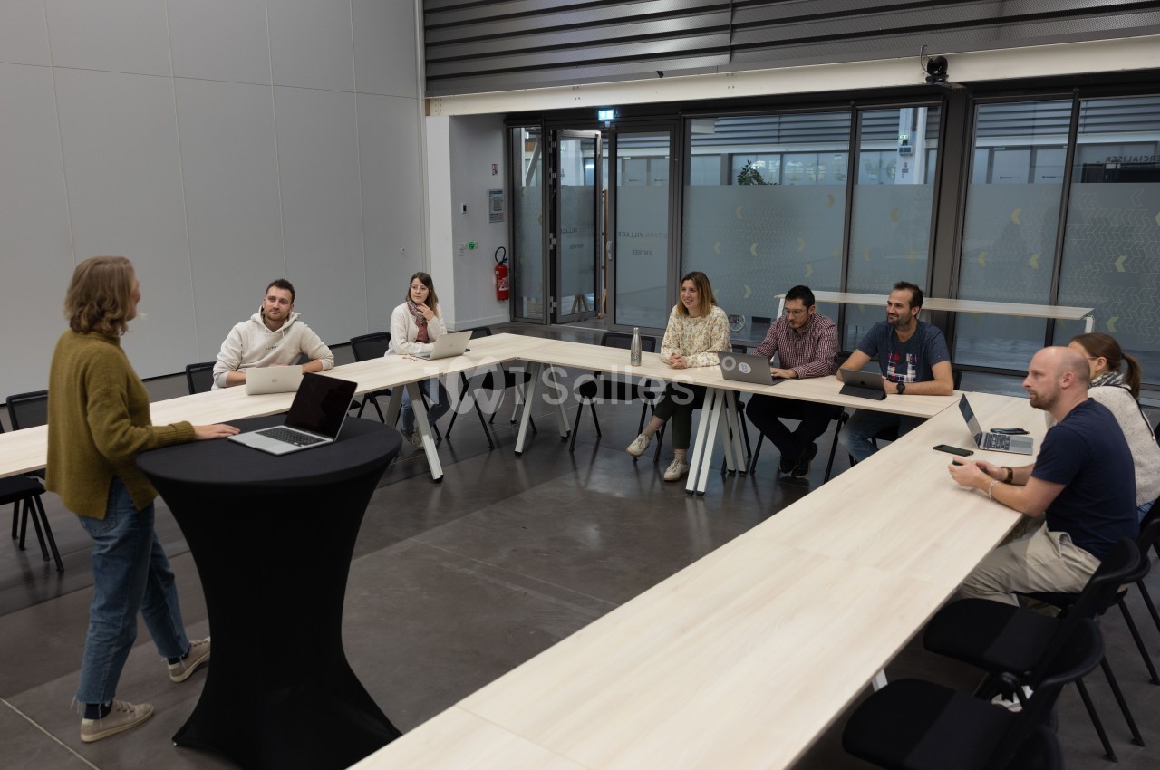 Une personne présente devant un groupe de collègues assis autour d'une table en U dans une salle de réunion moderne.