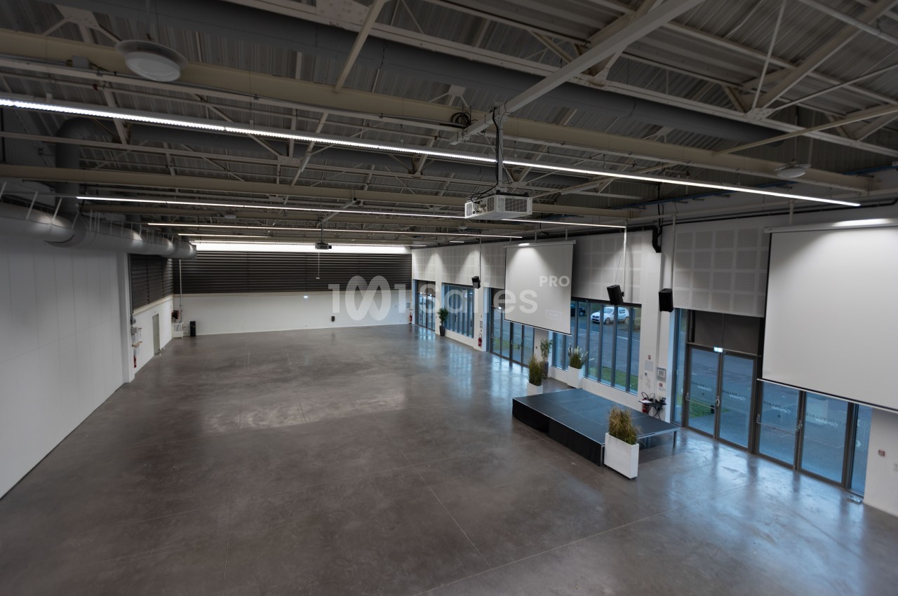 Salle vide avec sol en béton, grandes baies vitrées, murs blancs et plafond industriel avec éclairage suspendu.