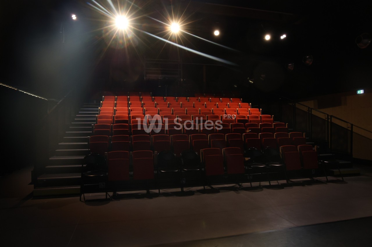 Salle de spectacle vide avec des rangées de sièges rouges et des projecteurs allumés éclairant la scène.