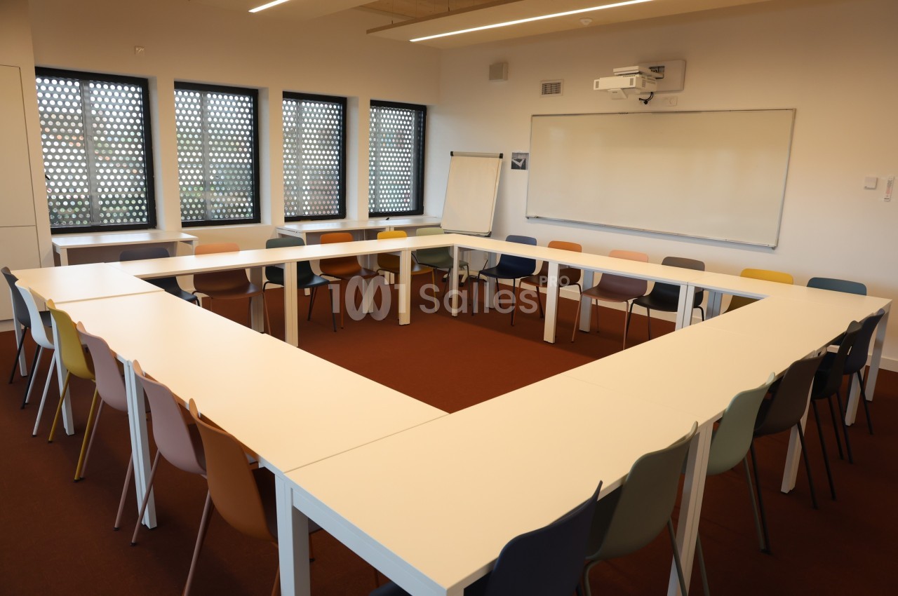 Salle de réunion avec tables disposées en U, chaises colorées, tableau blanc et paperboard près des fenêtres.