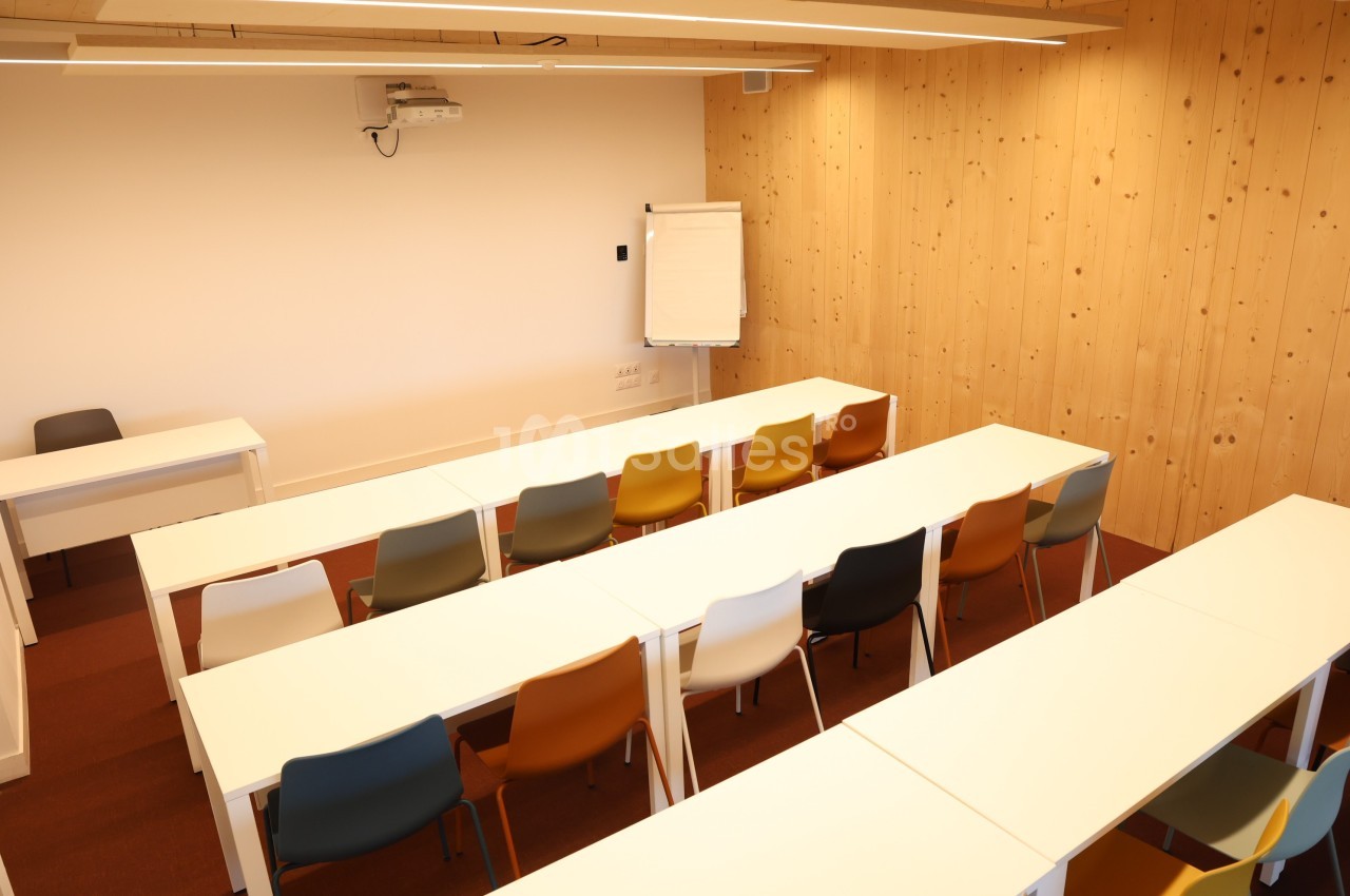 Salle de formation lumineuse avec des tables blanches alignées, des chaises colorées et un tableau de présentation.
