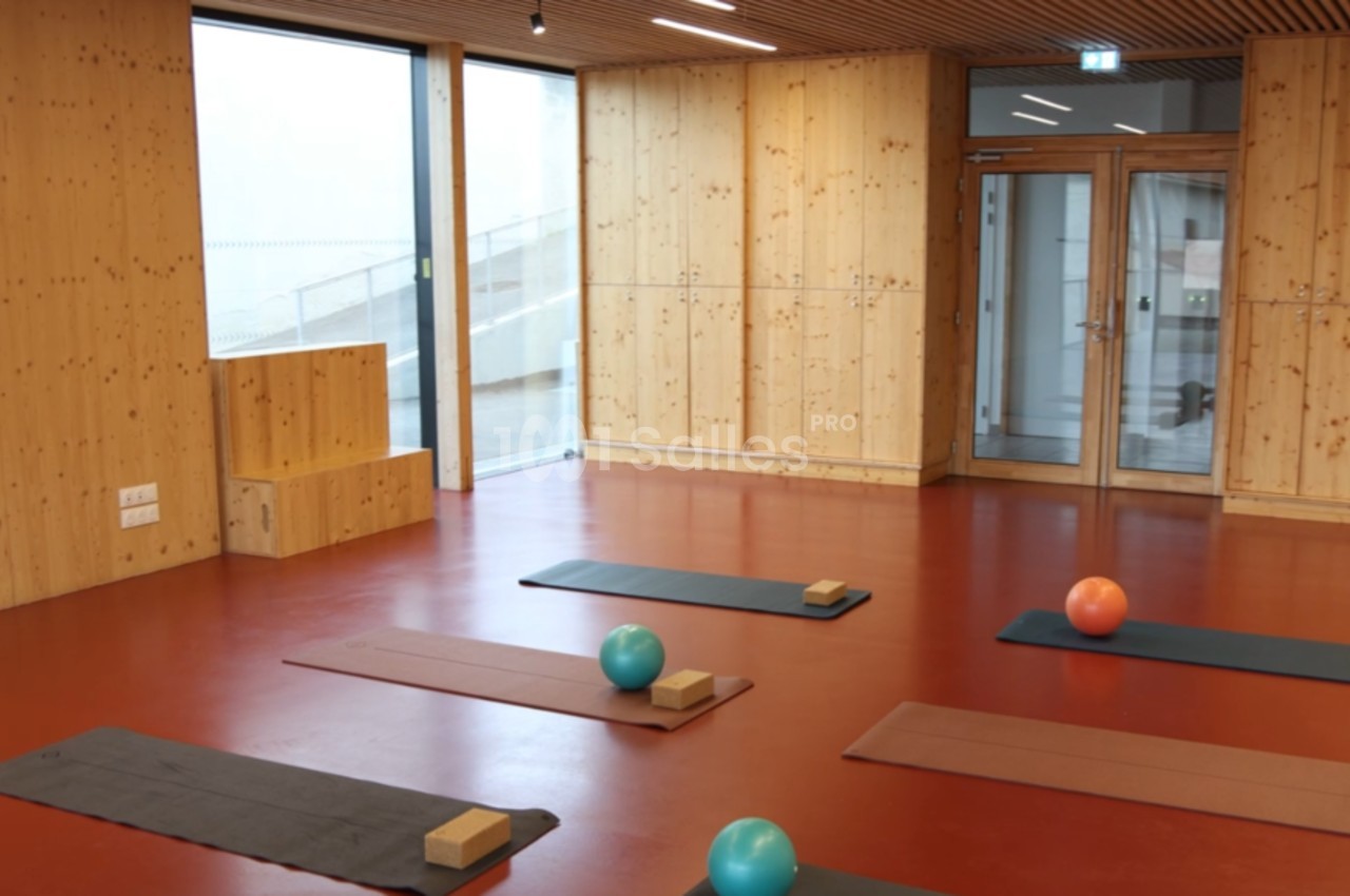 Salle lumineuse avec sol rouge, murs en bois, tapis de yoga, ballons et briques disposés pour une séance.