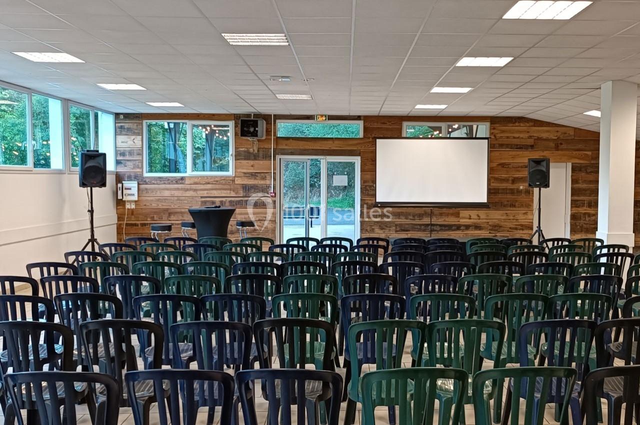 Salle de conférence avec des rangées de chaises en plastique, un écran de projection et des murs en bois.