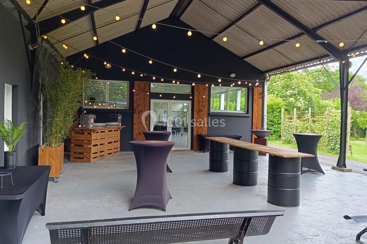 Terrasse couverte avec tables hautes, tonneaux en bois, guirlandes lumineuses et vue sur un espace vert.