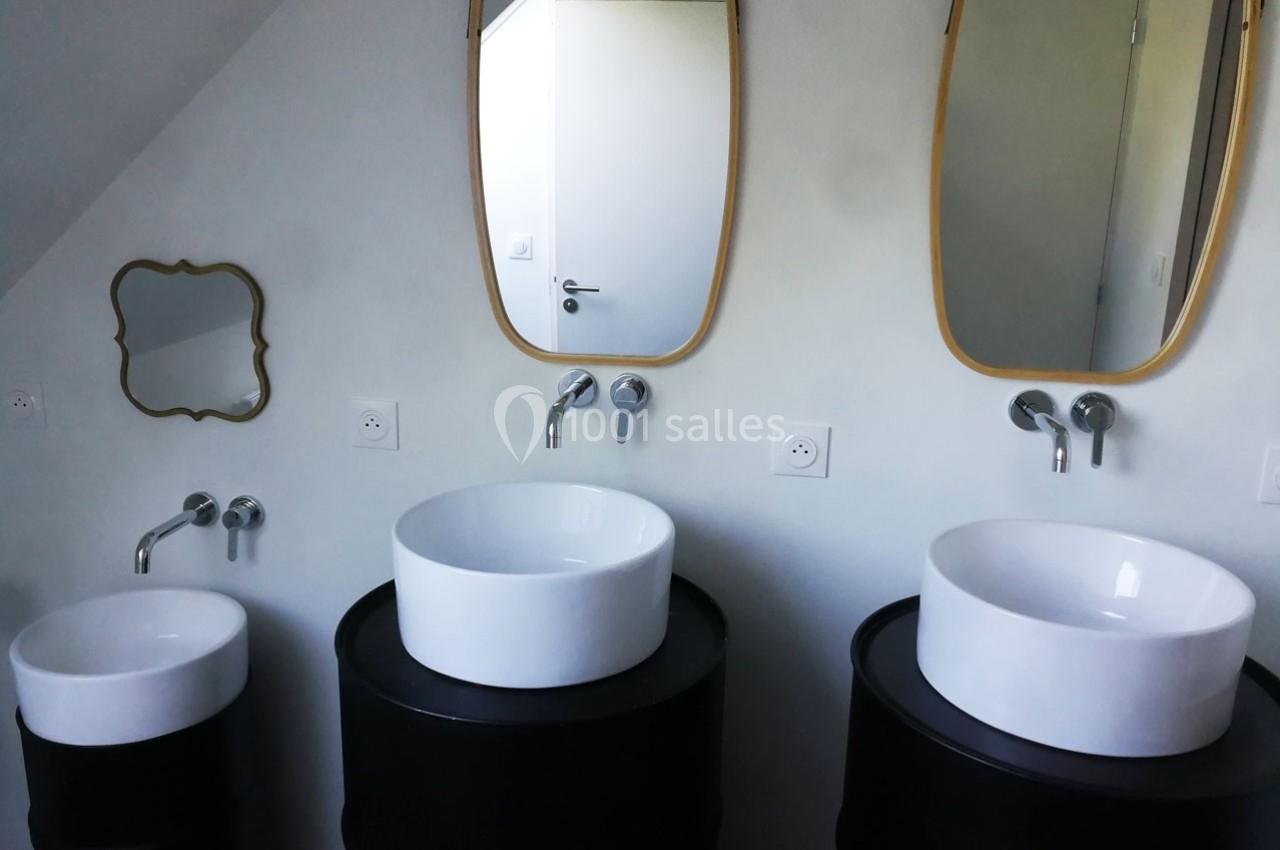 Trois lavabos blancs sur des supports noirs, avec miroirs ovales et mur blanc en arrière-plan.