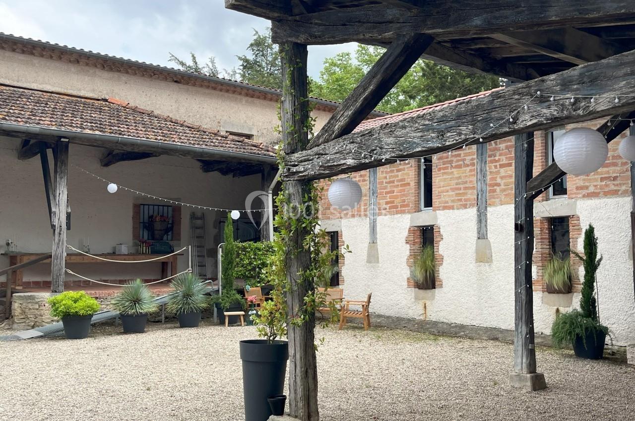 Cour intérieure avec gravier, pergola en bois, plantes en pots et lanternes suspendues sous un ciel nuageux.