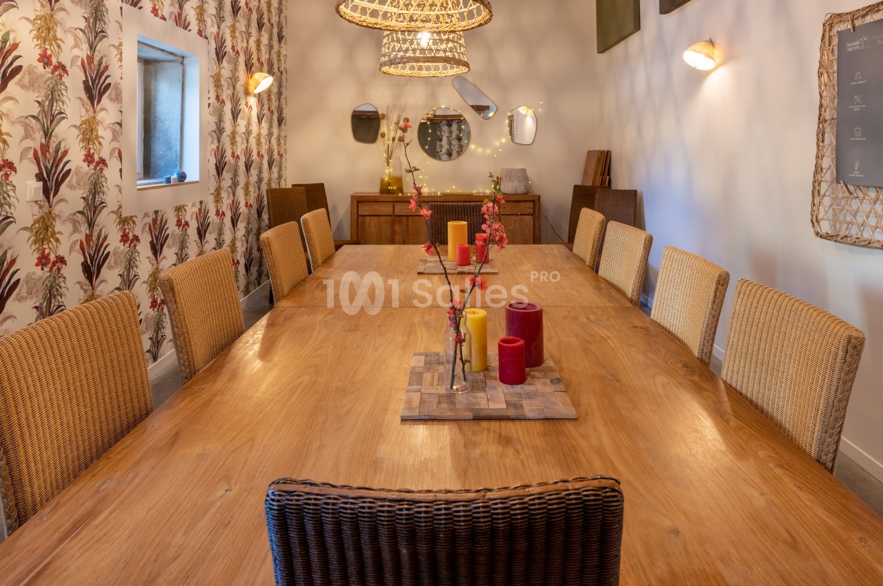 Grande table en bois entourée de chaises en osier, décorée de bougies et de branches, dans une salle à la lumière…