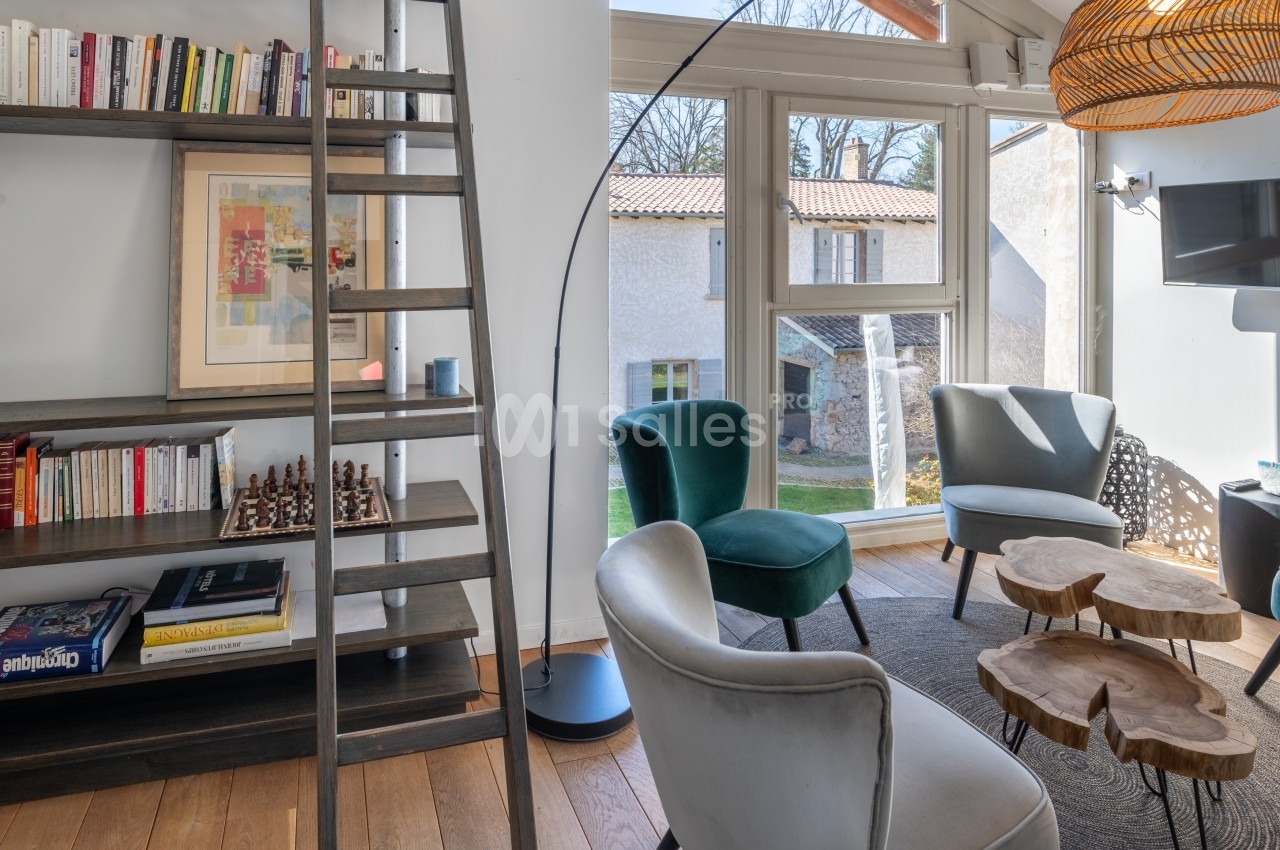 Salon lumineux avec fauteuils, table basse en bois, bibliothèque avec échelle et vue sur un jardin à travers une baie vitrée.