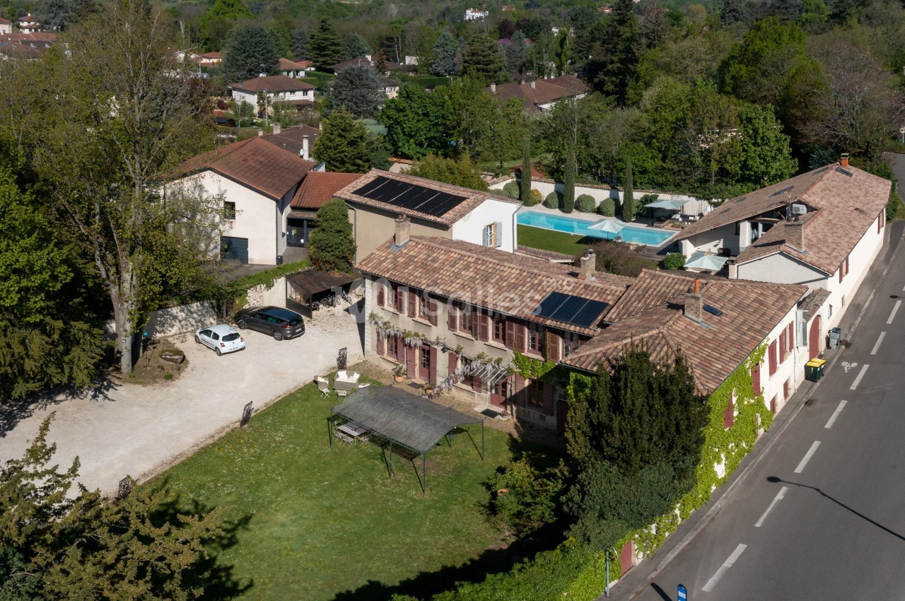 Vue aérienne d'une propriété avec plusieurs bâtiments, panneaux solaires sur les toits, piscine et végétation environnante.