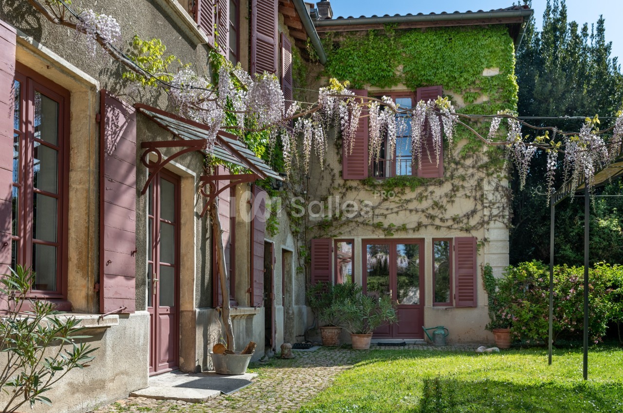 Façade d'une maison ancienne avec volets rouges, glycines en fleurs et jardin verdoyant.