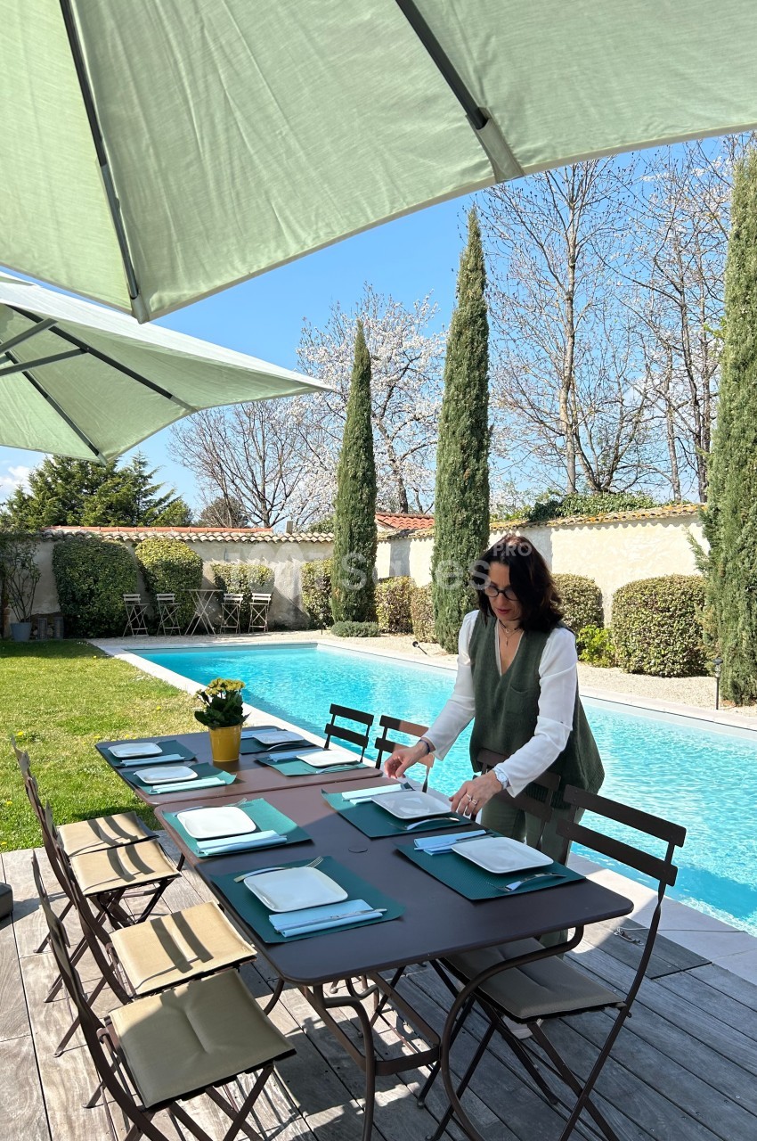 Une femme prépare une table extérieure dressée près d'une piscine, entourée de parasols et de végétation.