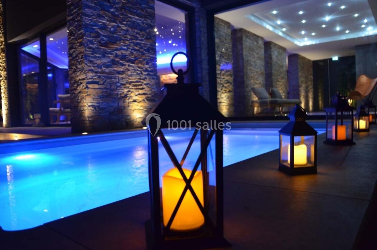 Piscine intérieure éclairée avec des lanternes à bougies sur le bord, entourée de murs en pierre et d'un plafond lumineux.