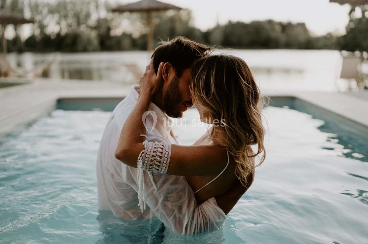 Un couple s'enlace dans une piscine, entouré d'un paysage naturel avec des arbres et un lac en arrière-plan.