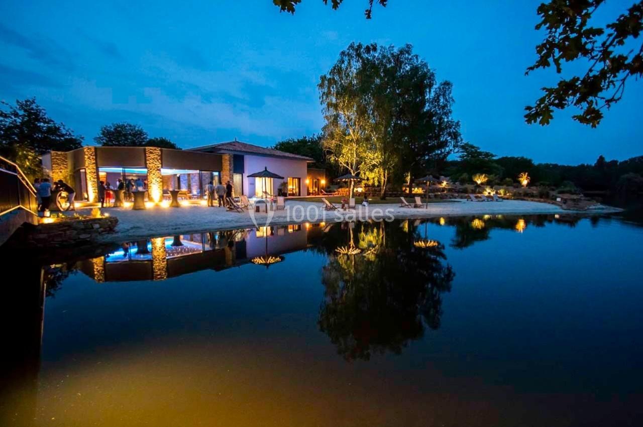 Vue nocturne d'un bâtiment éclairé au bord d'un étang calme, reflétant les lumières et les arbres environnants.