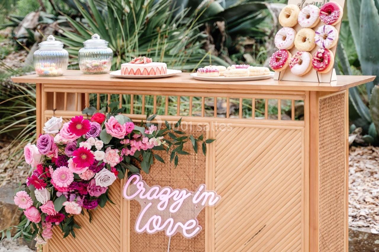 Table en bois décorée de fleurs roses, enseigne lumineuse ’Crazy in love’ et desserts variés, dont des donuts.