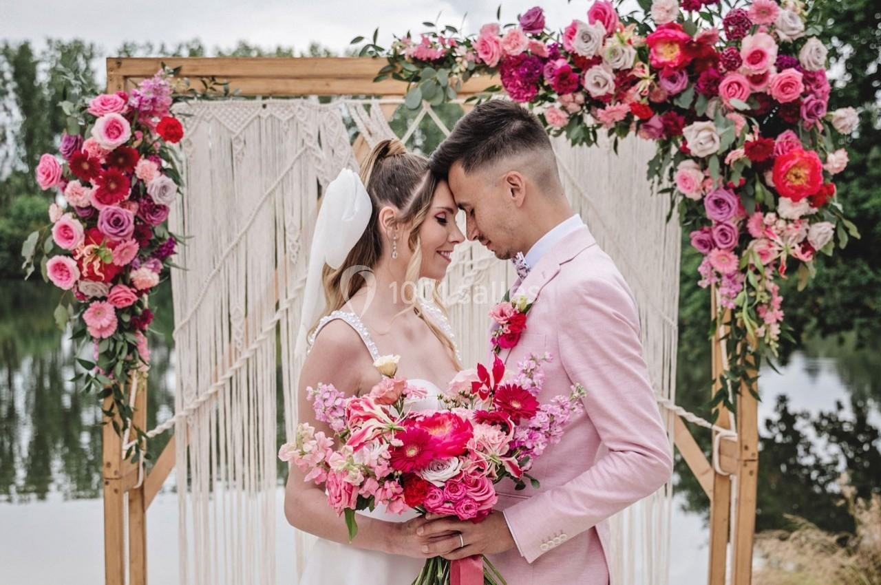 Un couple de mariés se tient face à face devant une arche décorée de fleurs roses près d'un plan d'eau.