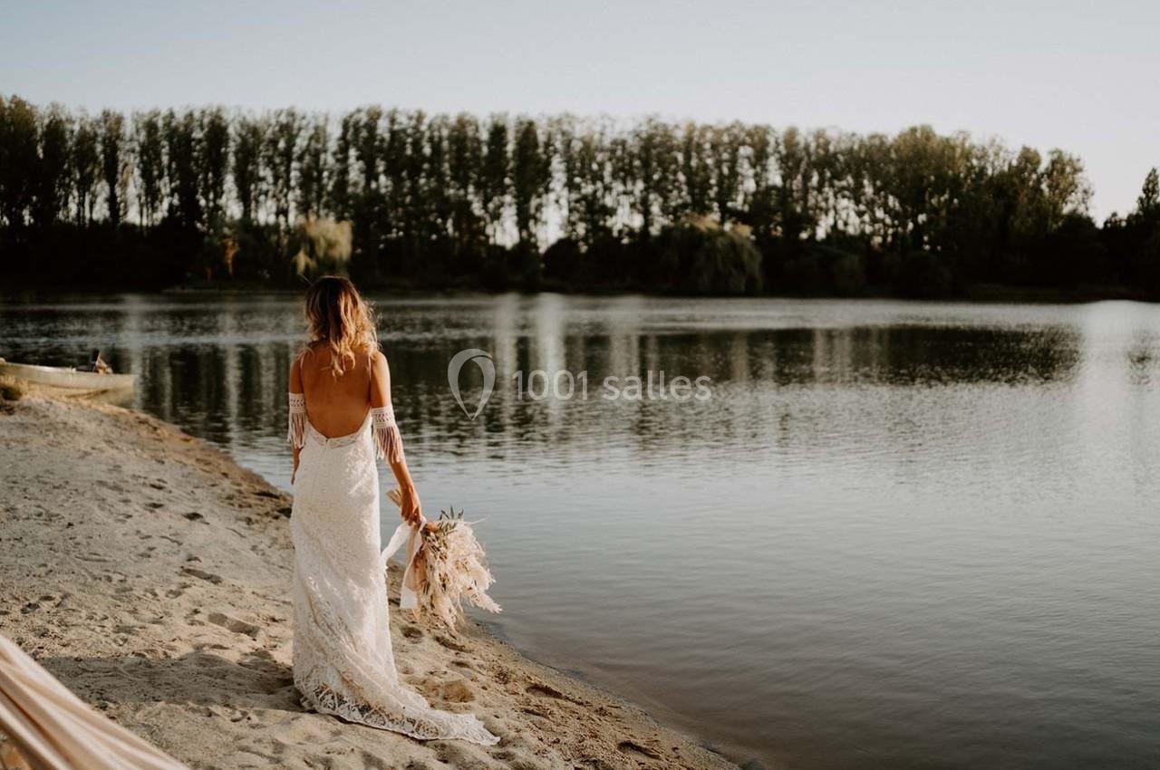 Une femme en robe de mariée marche sur une plage au bord d'un lac, tenant un bouquet de fleurs séchées.