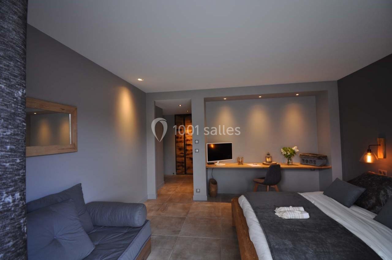 Chambre moderne avec lit double, bureau en bois, fauteuil gris et éclairage tamisé dans un décor sobre et épuré.