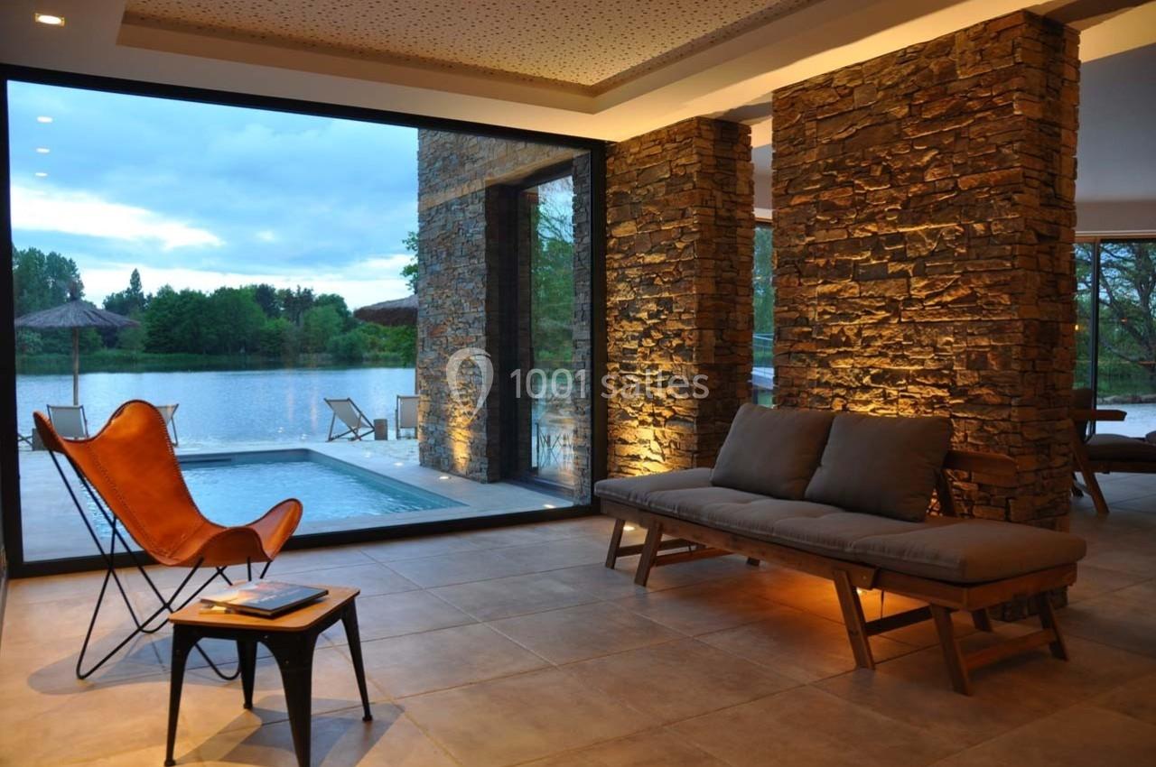 Salon moderne avec murs en pierre, grandes baies vitrées donnant sur un lac, fauteuil en cuir et piscine extérieure.