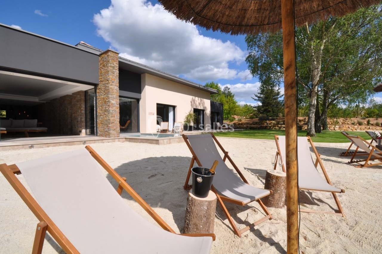 Transats en bois et toile beige disposés sur du sable devant une maison moderne avec terrasse et jardin arboré.